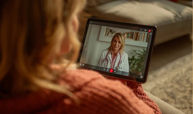 Paciente en un sofá sosteniendo una tableta durante una videoconsulta de telesalud con una doctora sonriente que lleva un estetoscopio